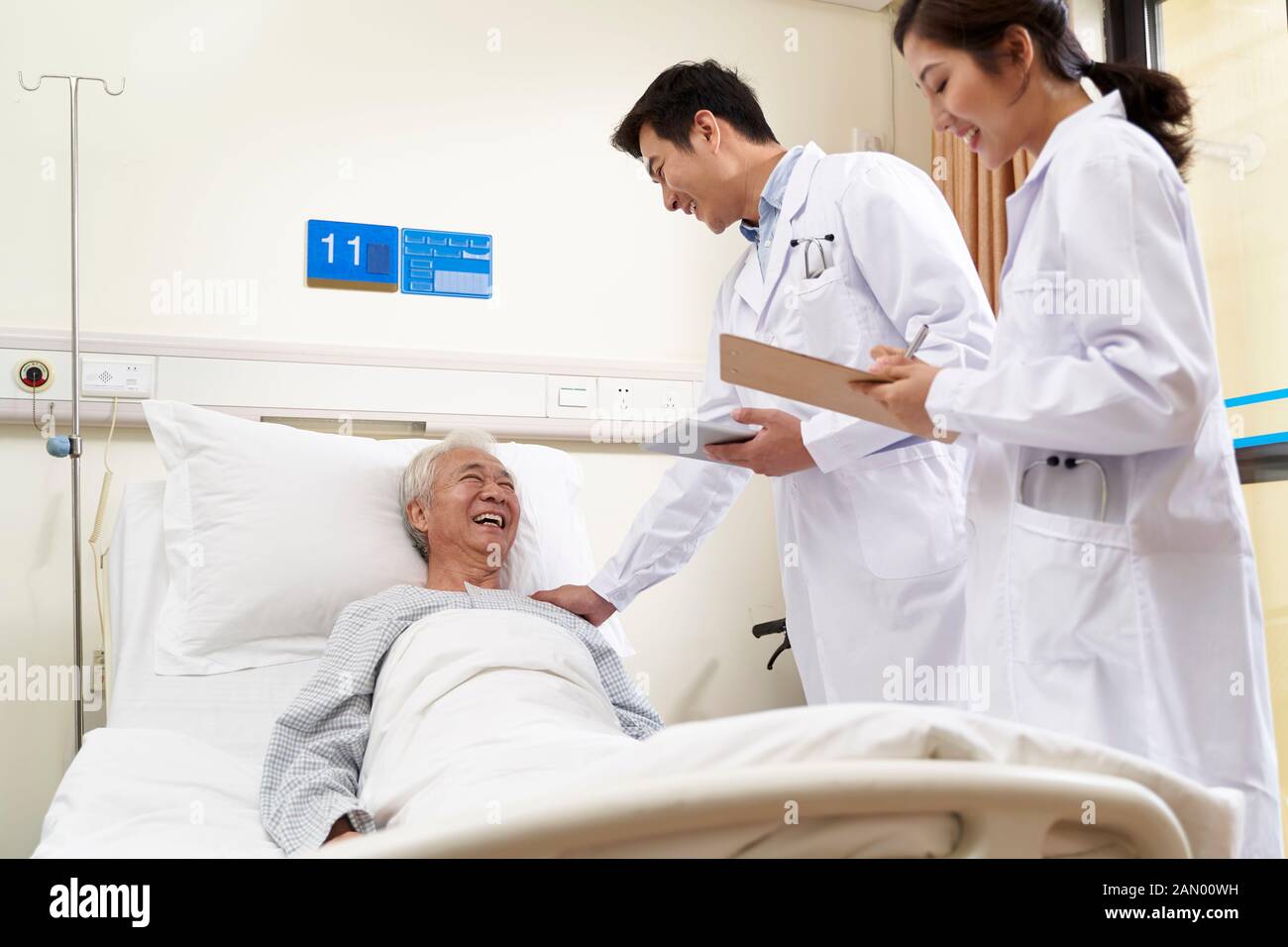 Zwei junge, freundliche asiatische Ärzte sprechen mit einem männlichen leitenden asiatischen Patienten am Krankenbett Stockfoto