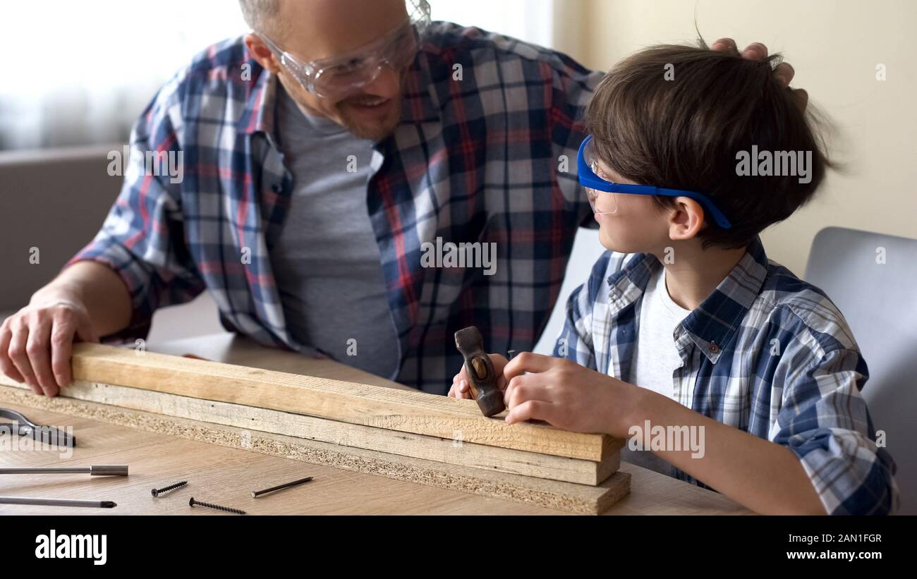 Niedlicher kleiner Junge, der Nagel in Holzpflaster hämmert, Vater lobt sein Kind, Stütze Stockfoto