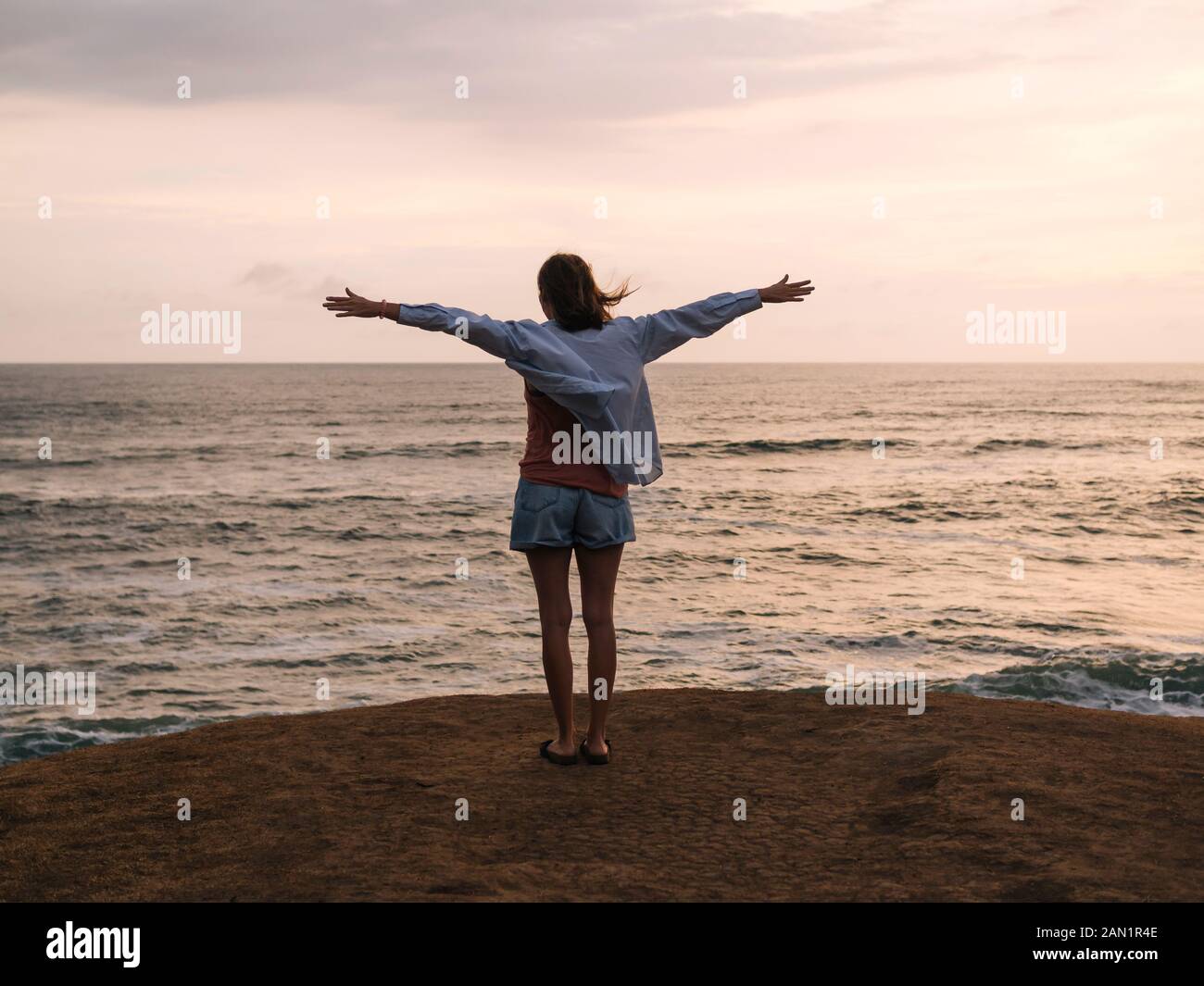 Frau mit erhobenen Armen, die bei Sonnenuntergang am Meer steht Stockfoto