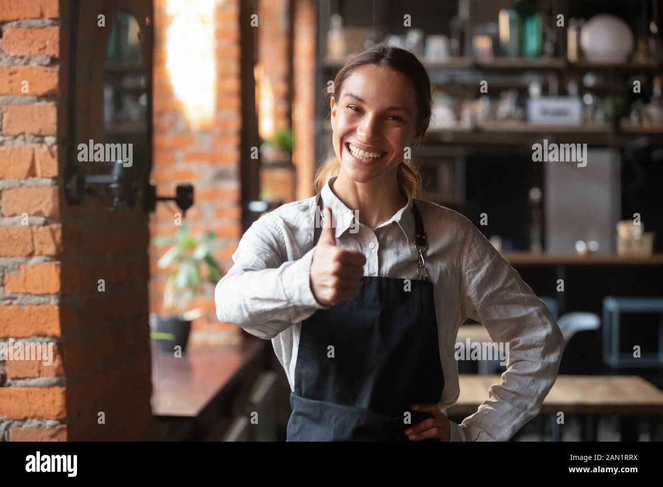 Lächelnde Kellnerin in der Schürze zeigt Daumen nach oben und empfiehlt Platz Stockfoto
