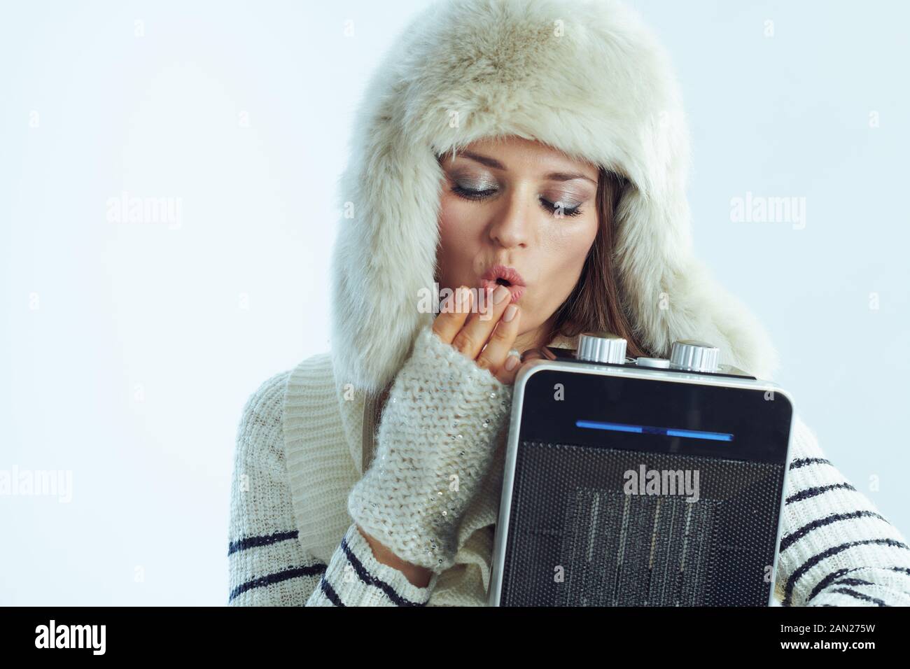 Gefrorene, stylische 40 Jahre alte Frau in weiß gestreifter Pullover, Schal- und Ohrenklappenhut, die tragbare elektrische Heizung hält und im Winter atmet l Stockfoto