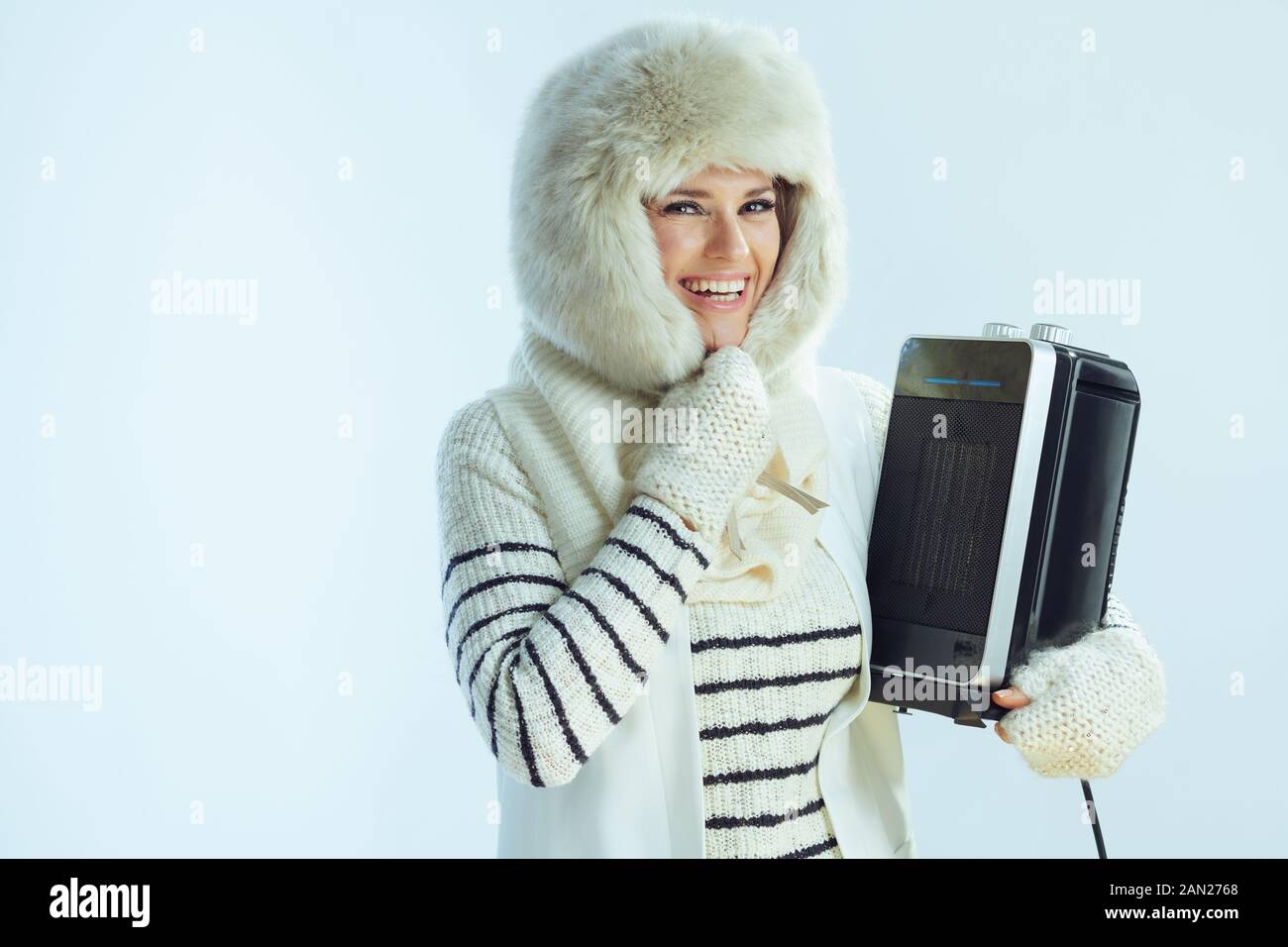 Porträt einer lächelnden modernen Frau im mittleren Alter in weiß gestreiften Pullover, Schal und Ohrenklappen Hut mit tragbarer Elektroheizung isoliert auf Winterhöhe Stockfoto