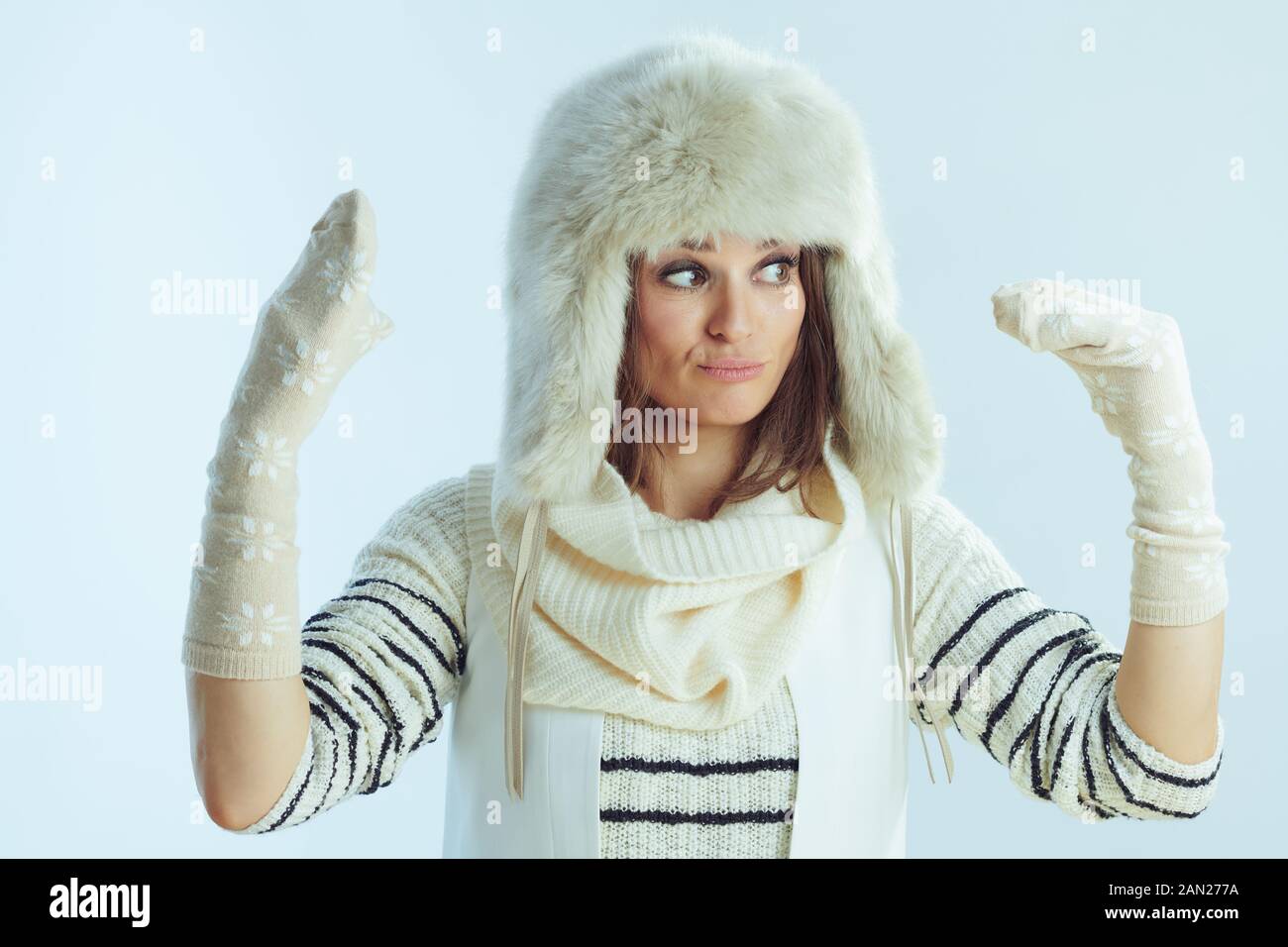 Lustige, elegante 40 Jahre alte Frau in weiß gestreiftem Pullover, Schal und Ohrenklappenhut, die mit Wintersocken auf winterhellblauem Hintergrund spielt. Stockfoto