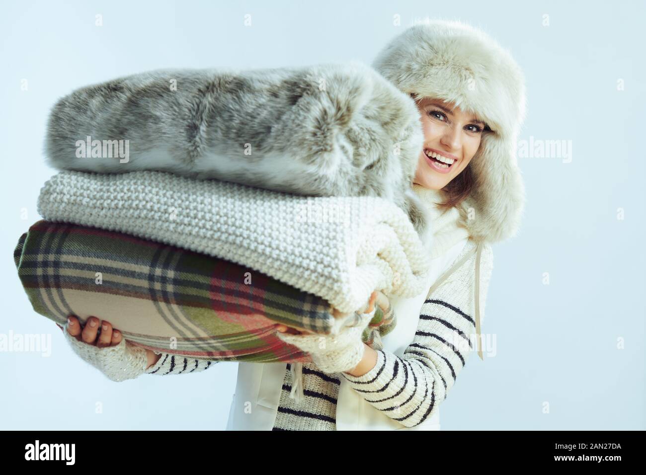 Porträt einer fröhlichen eleganten Frau in weiß gestreiftem Pullover, Schal und Ohrenklappenhut, die einen Stapel Decken vor winterhellblauem Hintergrund hält. Stockfoto