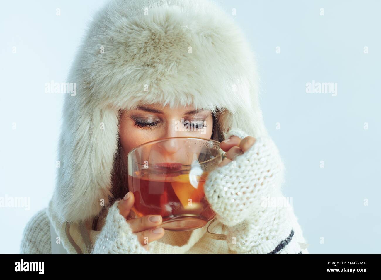 Elegante Frau in weiß gestreiftem Pullover, Schal und Ohrenklappen hat Trinkbecher mit heißem Tee mit Zitrone und Honig auf winterhellblauem Hintergrund. Stockfoto