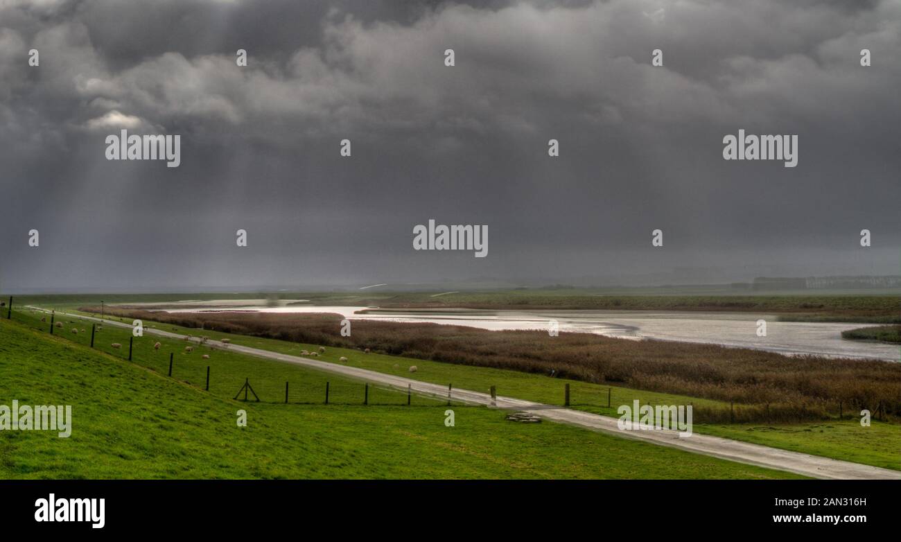 Sonnenstrahlen in einen dunklen Himmel über die Küstenlandschaft im Norden der Niederlande; ein Deich und Feuchtgebiete Stockfoto