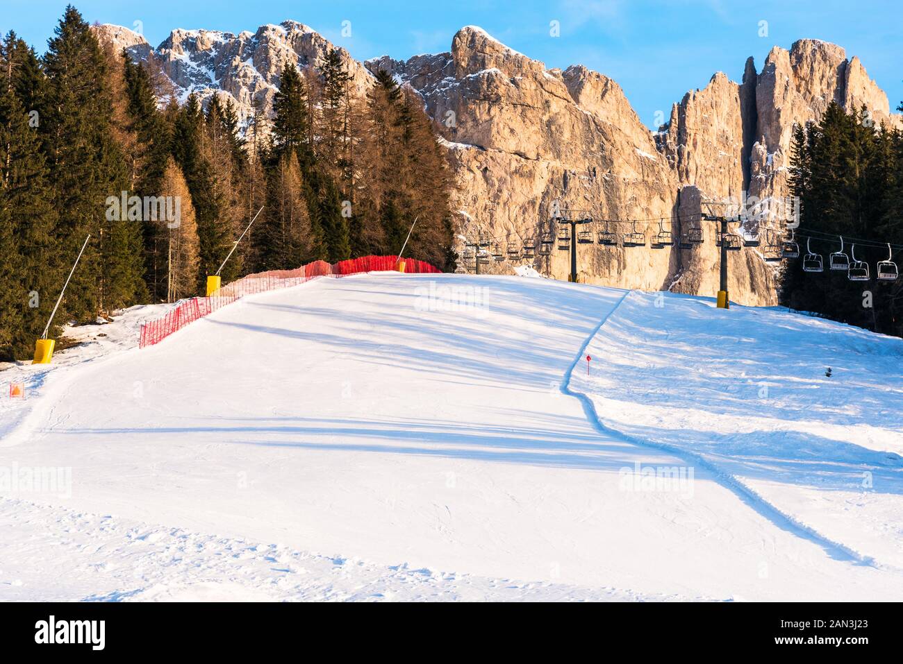 Einsame Piste mit einem Sessellift im Hintergrund am Fuße der hoch aufragenden felsigen Gipfeln der Dolomiten auf einem klaren Winter am Nachmittag Stockfoto