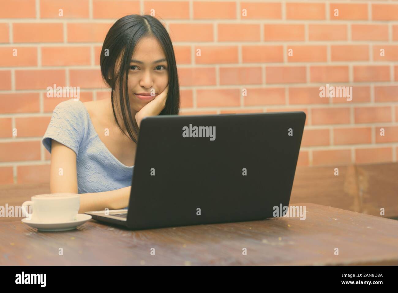Junge schöne asiatische Jugendmädchen ruhenden Kinn auf Hand mit Laptop und Cappuccino auf hölzernen Tisch gegen die Wand Stockfoto