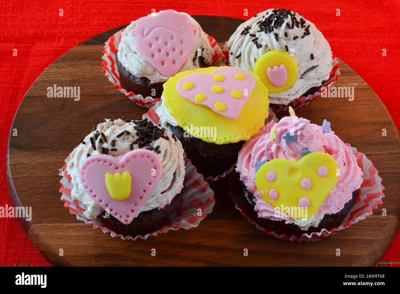 Liebe Muffins, bunten Cupcakes mit Marzipan Herzen, auf Holz- runde Untertasse über den roten Tischdecke serviert dekoriert, Seitenansicht Stockfoto