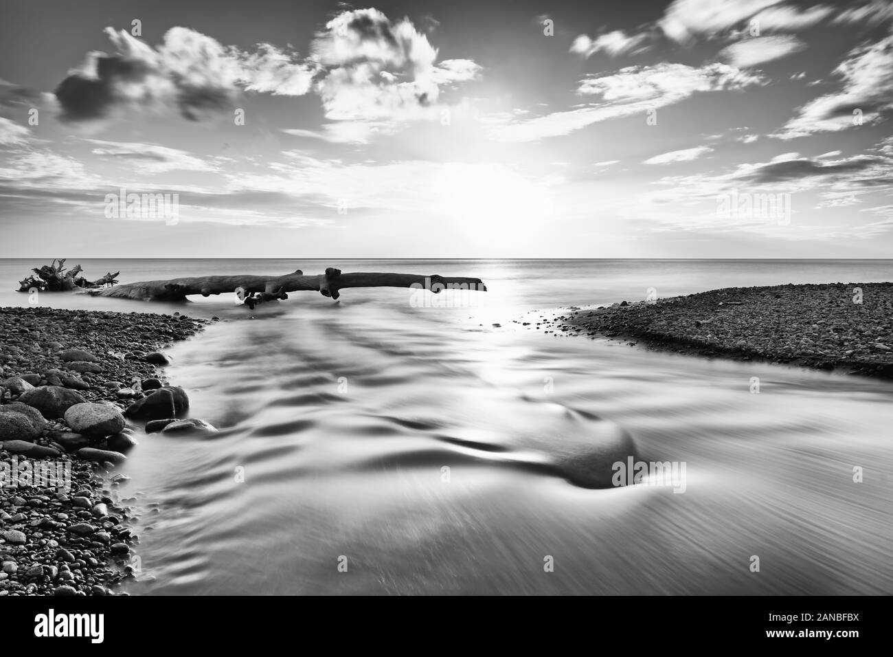 Malerischen Sonnenuntergang auf einem Stein Strand, Blick entlang einem kleinen Fluss, der ins Meer fließt, angeschwemmte Baum vor dem Horizont, Wellen, lange Zeit exposur Stockfoto