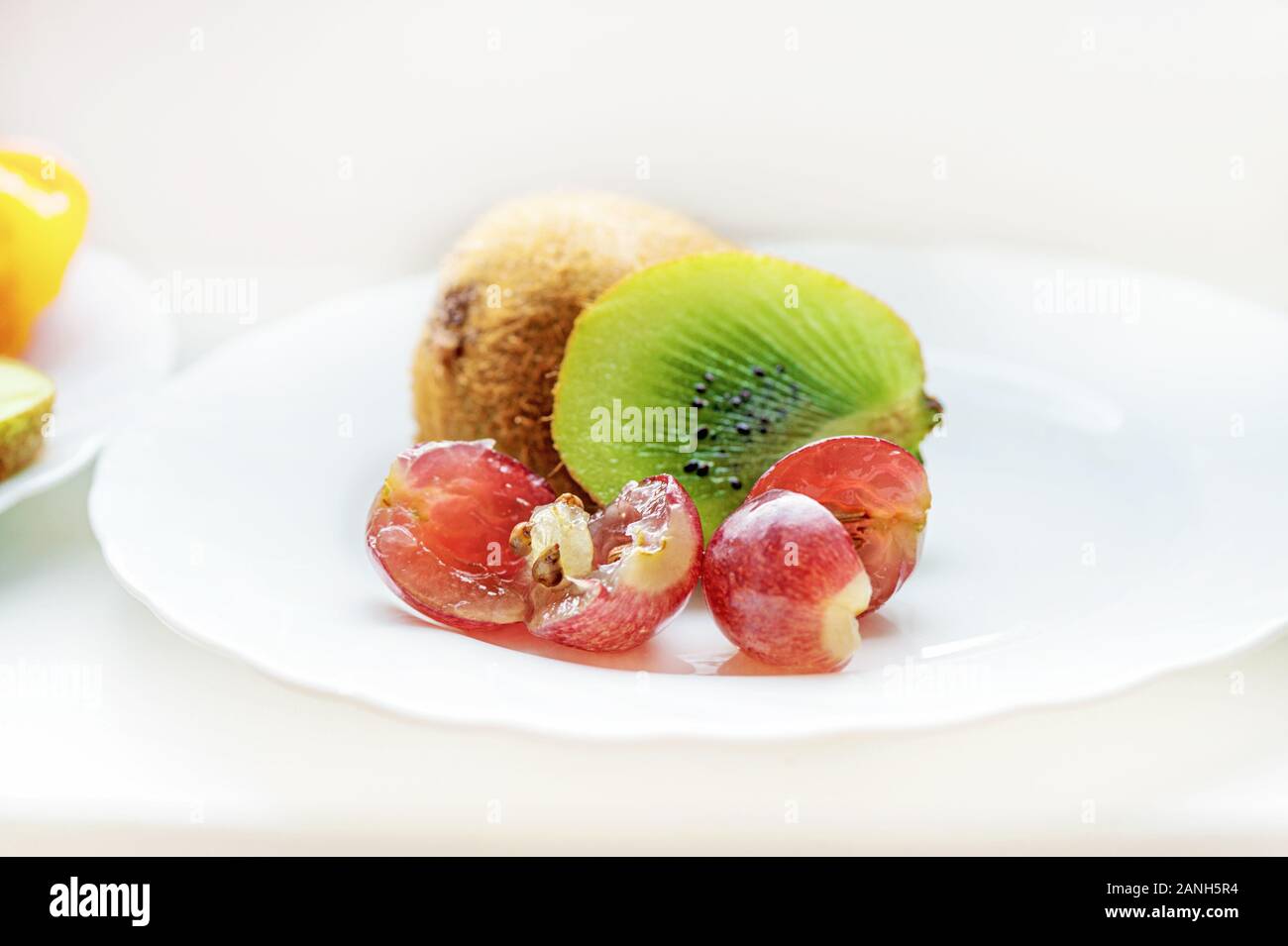 Auf der Platte ist die Hälfte eines sehr reife, saftige grüne Kiwi mit Trauben auf einem weißen Teller. Close Up. Licht monochromen Hintergrund. Stockfoto