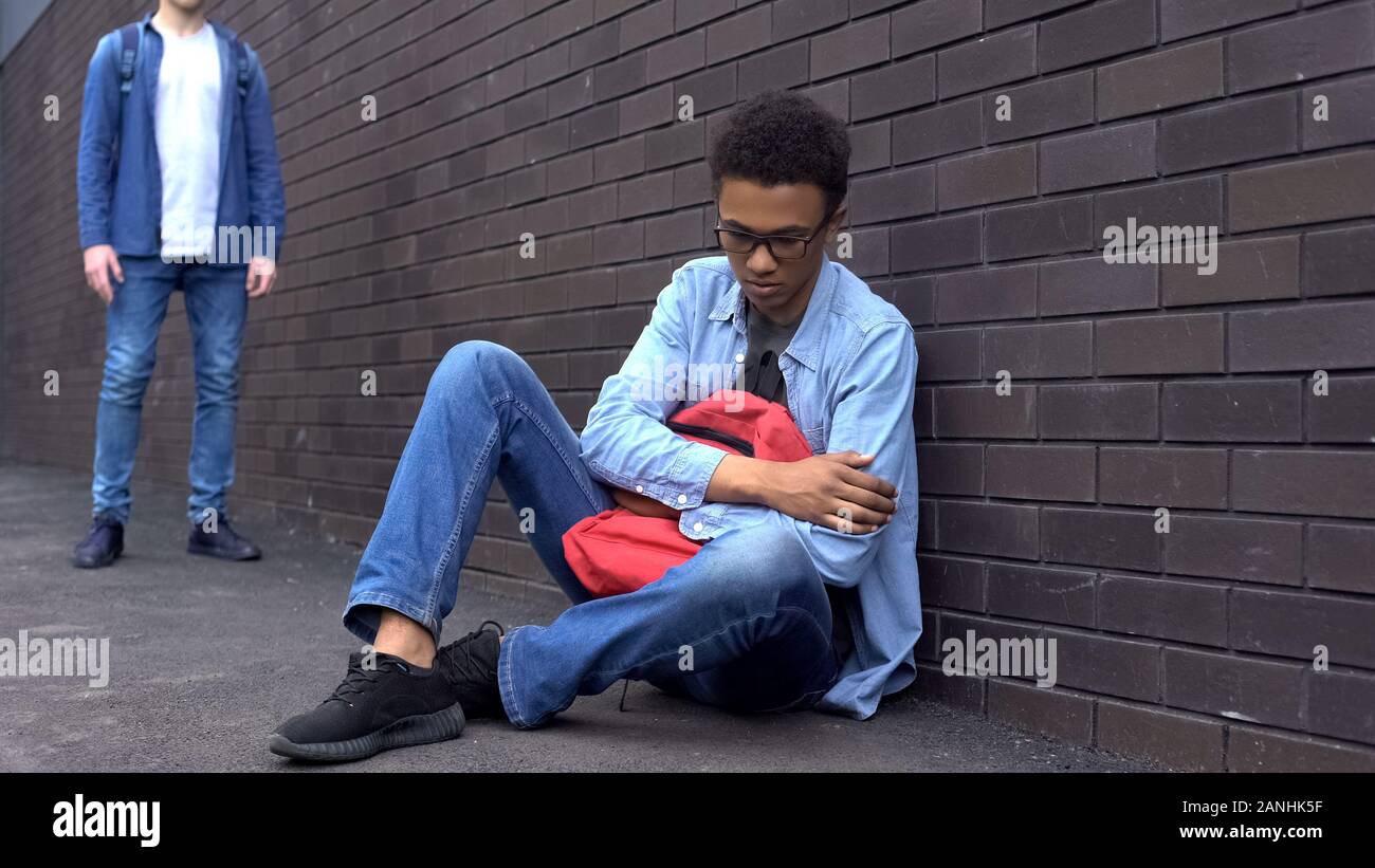 Tyrannisiert afro-amerikanischen Schüler sitzen auf dem Boden, Angst der Täter hinter Stockfoto