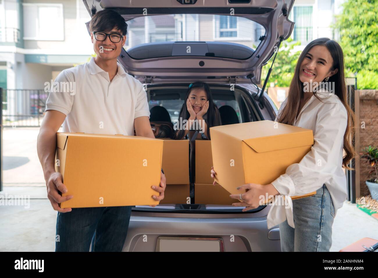 Gerne asiatische Familie mit Vater und Mutter steht in der Nähe von Auto mit Kartons und ihre Tochter lächelnd im Auto am Haus Garage. Beweglicher Tag. Stockfoto