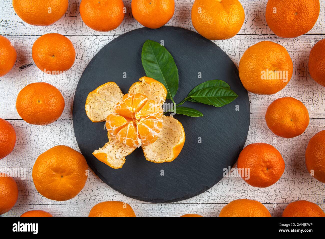 Geöffnet Mandarine mit Blättern auf runde, schwarze Platte mit den ganzen hellen orange Mandarinen auf Risse weiße Holztisch umgeben. Früchte haben natürliche Stockfoto