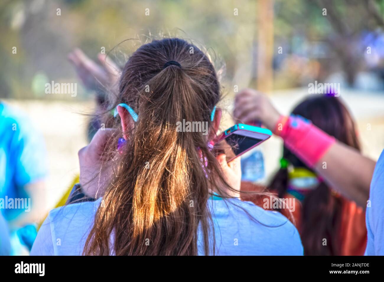 Holi Color Festival - Mädchen mit Pony-Schwanz und türkisfarbenen Sonnenbrillen und hellfarbigem Telefon in der Hand läuft durch eine Farbstation - nicht erkennbar mit Stockfoto