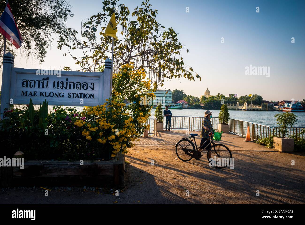 Mae Klong/Thailand-Man trägt eine Maske, die neben seinem Fahrrad am Ende der Eisenbahnlinie von Mae Klong läuft, mit Fluss im Hintergrund bei Sonnenuntergang. Stockfoto