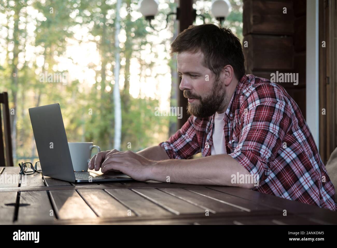 Bärtiger Mann arbeitet mit einem Laptop auf der Terrasse im Garten, vor dem Hintergrund der Bäume. Stockfoto