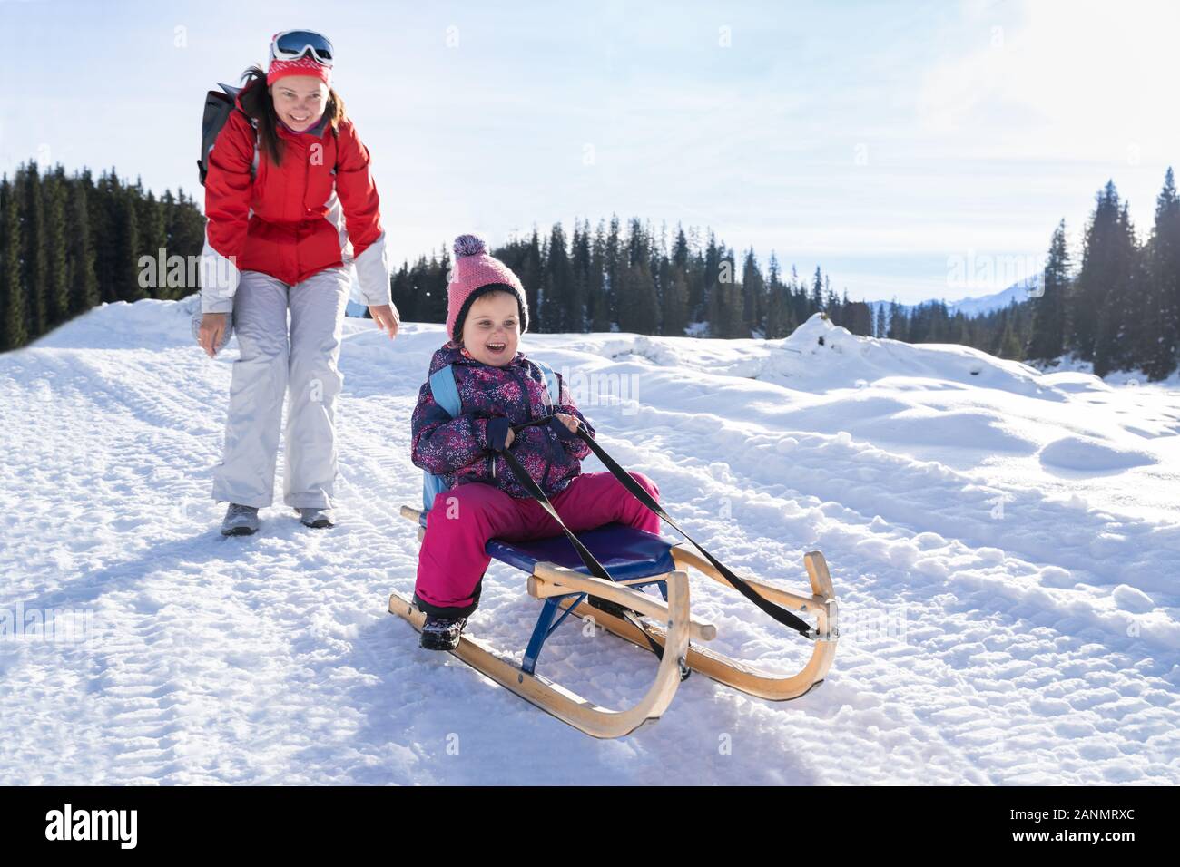 Adorable Girl Rodeln genießen, während ihre Mutter sie Schlitten gegen Pinienwald im Winter Stockfoto