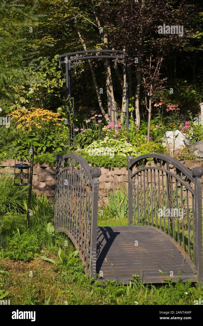 Alte schwarze schmiedeeiserne Metallbrücke und erhöhte Steingrenze mit gelben Rudbeckias, Hosta Pflanzen und lila Echinacea purpurea - Kegelblumen Stockfoto