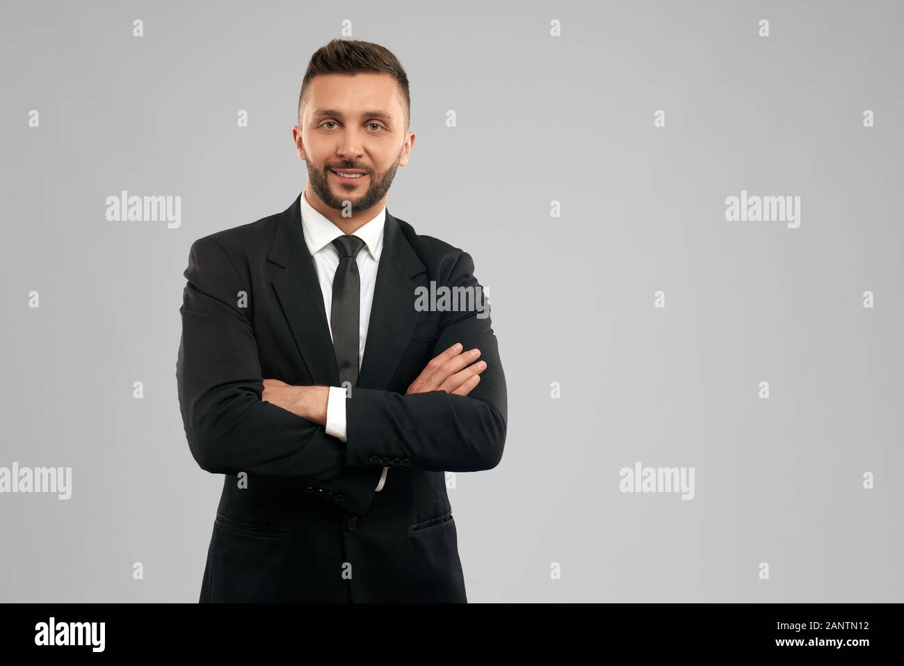 Vorderansicht des jungen lächelnden Geschäftsmannes in schwarzem Formanzug, der mit gekreuzten Händen posiert. Erfolgreicher brünette Arbeiter, der auf grauem Hintergrund steht und die Kamera betrachtet. Unternehmenskonzept. Stockfoto