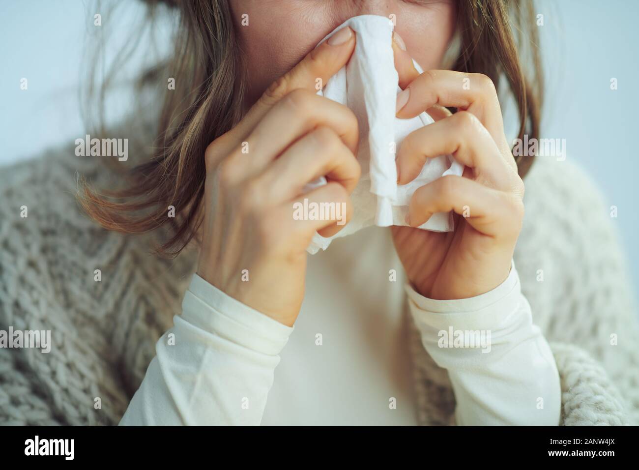 Nahaufnahme auf Kranke elegante Frau in Rollkragen Pullover und Strickjacke abwischen Nase mit Serviette auf Winter hellblau hinterlegt. Stockfoto