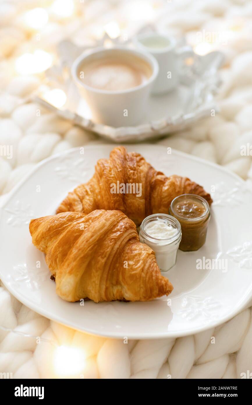 Frische Croissants mit Marmeladen und Americano mit Milch auf gestrickter weißer Wolldecke und leuchtenden Girlanden. Gemütlicher Wintermorgen zu Hause. Skandinavisches Schlafzimmer. Stockfoto