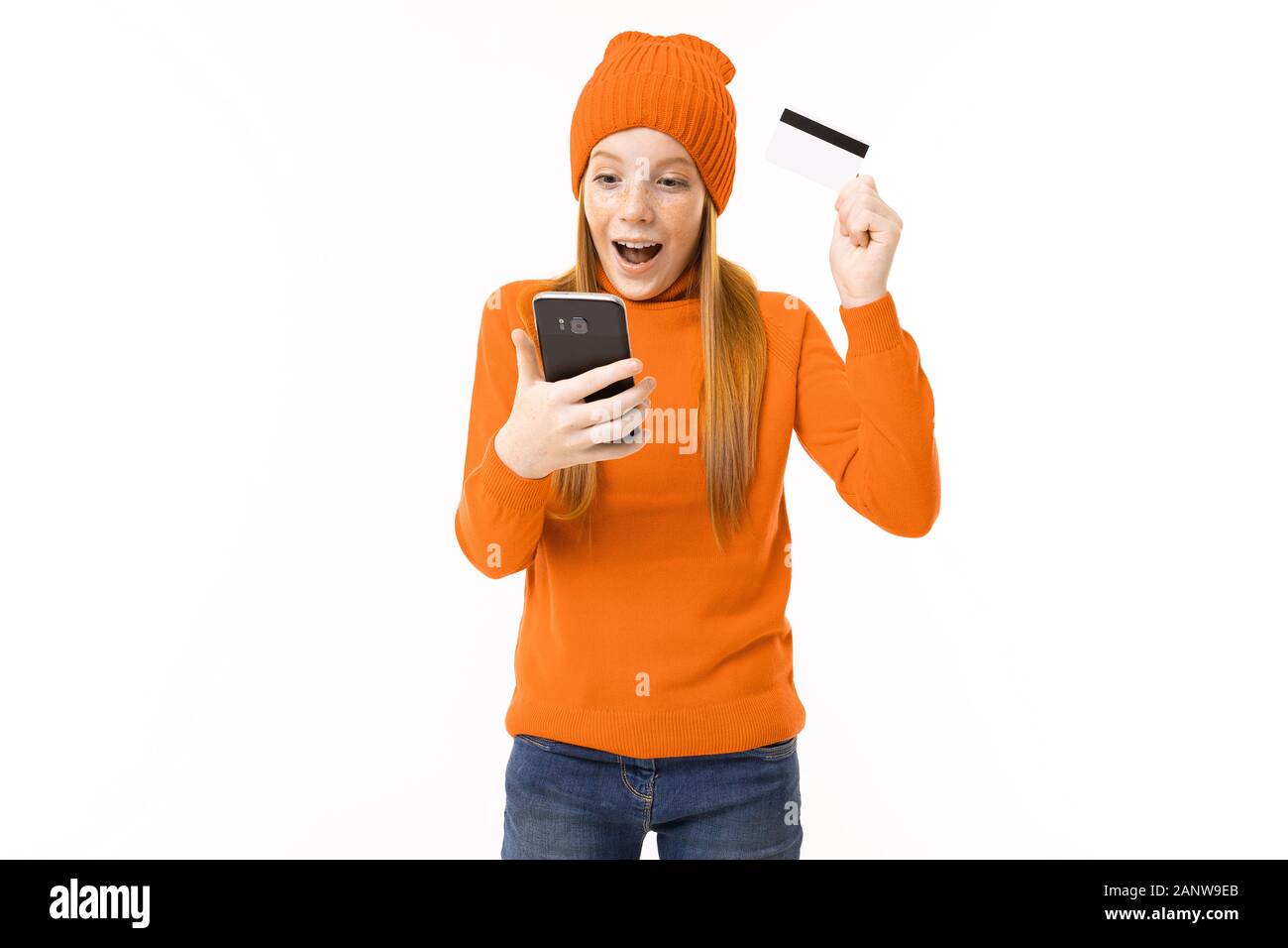 Fröhliches Mädchen mit roten Haaren, orangefarbenem Hoodie und Hut mit Kreditkarte und Telefon isoliert auf weißem Hintergrund Stockfoto