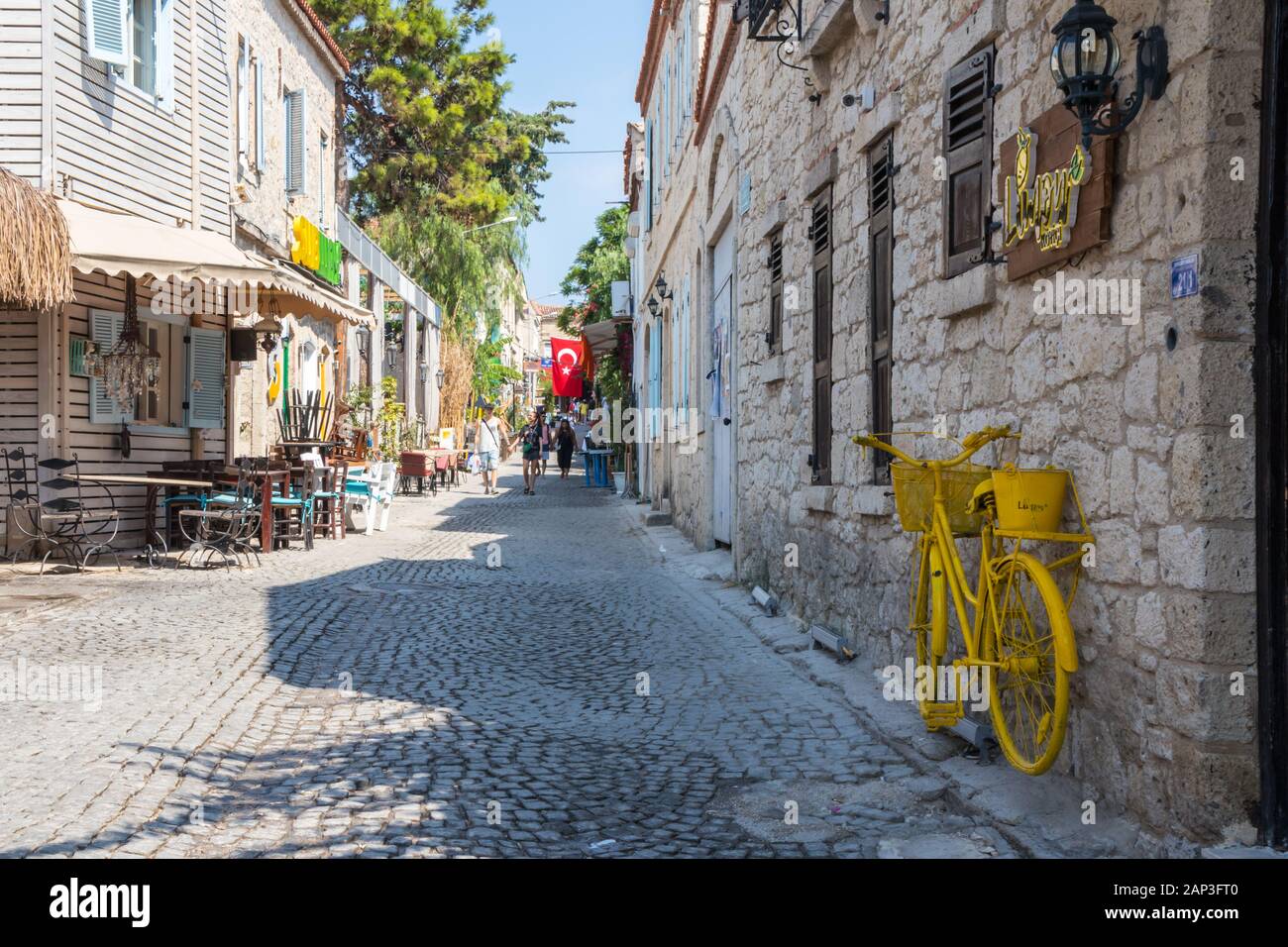 Alacati, Türkei - September 2019: Menschen gehen auf einer gepflasterten Straße und gelben Fahrrad. Viele Touristen besuchen die Stadt. Stockfoto