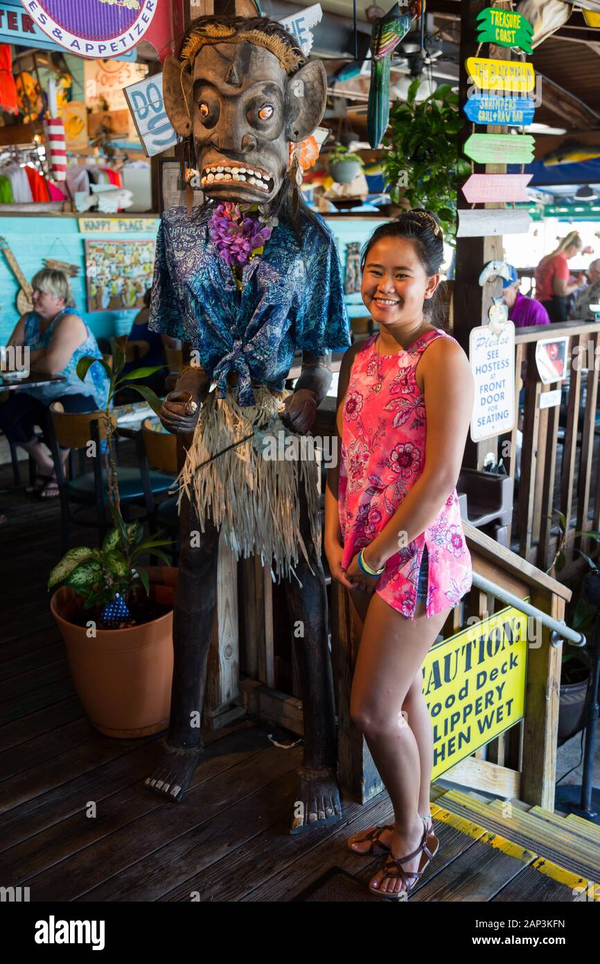 Ein schönes Teenager-Mädchen steht neben einem hässlichen, unheimlichen Tiki-Idol in farbenfrohem Shrimper's Grill and Raw Bar in Port Salerno, Florida, USA. Stockfoto