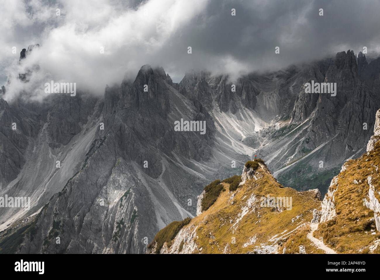 Frau in der roten Jacke stehend auf einem Bergrücken, hinter Gipfeln und spitzen Felsen, dramatische Wolken, Cimon di Croda Liscia und Cadini Gruppe, Auronzo di Stockfoto