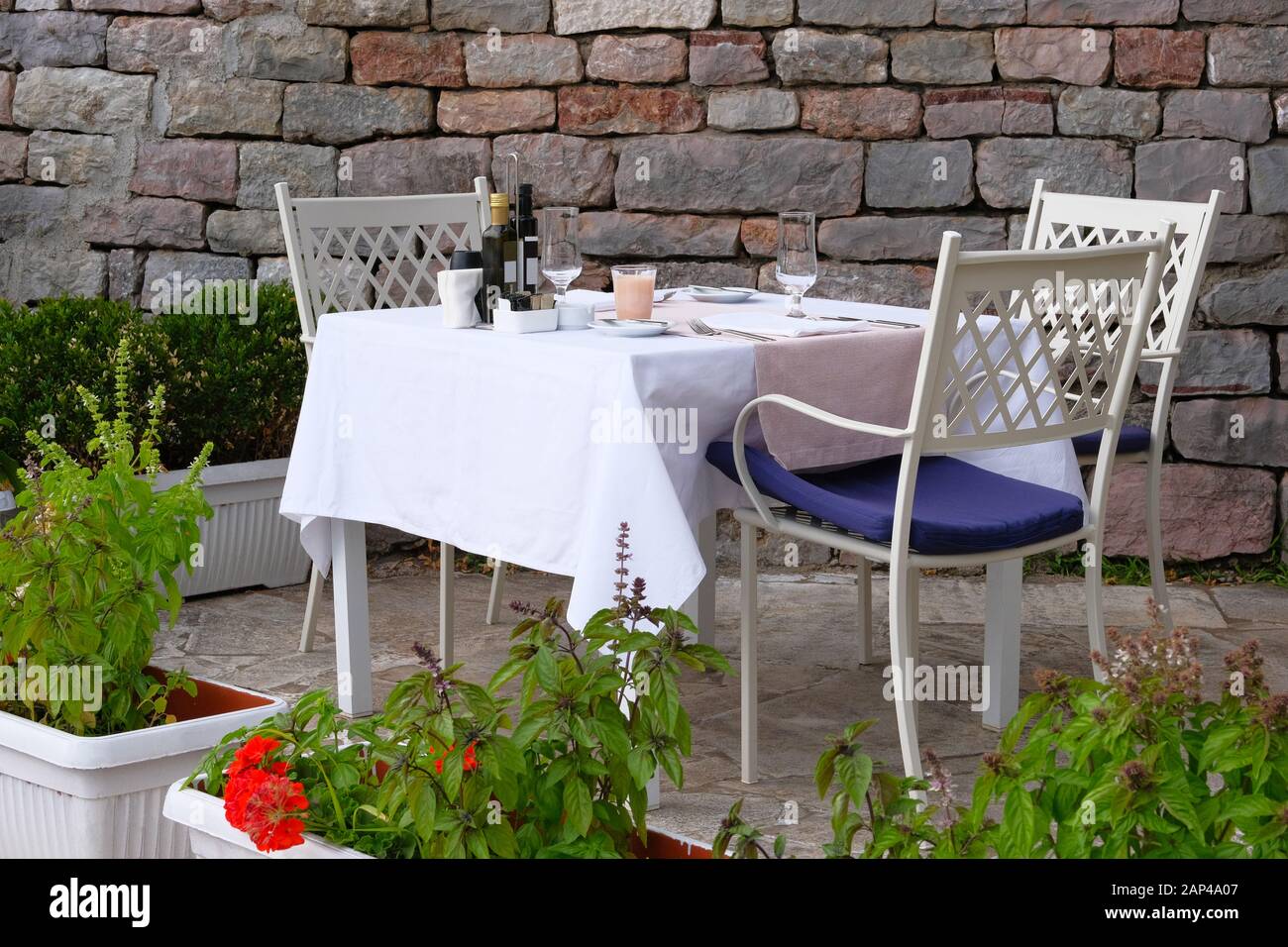 Tisch mit weißen Tischdecken, Besteck und Weingläsern auf der Sommerterrasse des Restaurants. Wand aus altem, strukturiertem Stein im Hintergrund. Stockfoto
