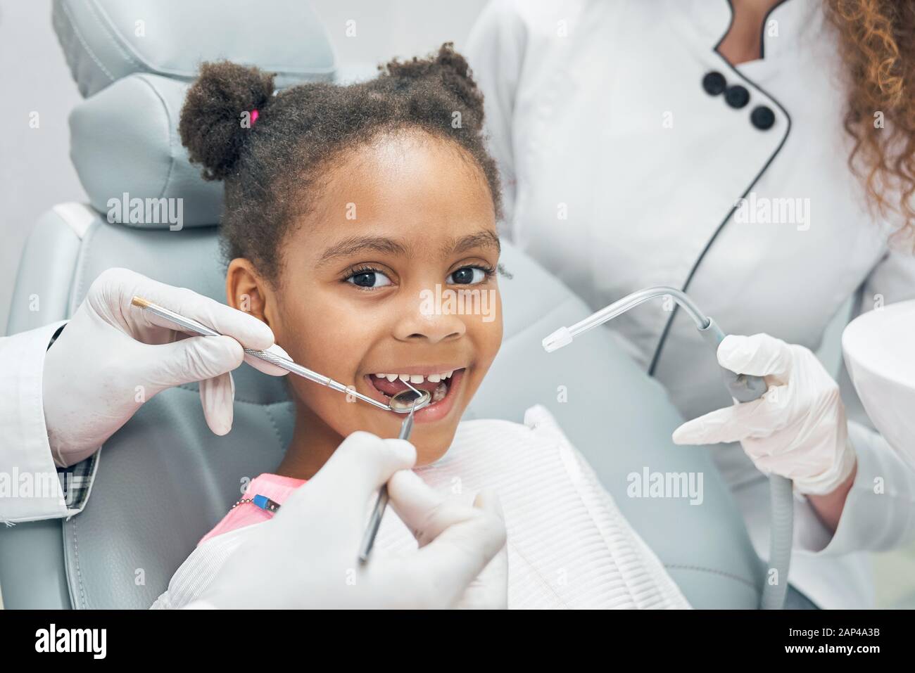 Glückliches afro-amerikanisches Mädchen, das im Stomatologenstuhl mit offenem Mund sitzt, während der professionelle Zahnarzt regelmäßig die Zähne mit einer Zahnsonde und einem Spiegel überprüft. Krankenschwester hilft. Stockfoto