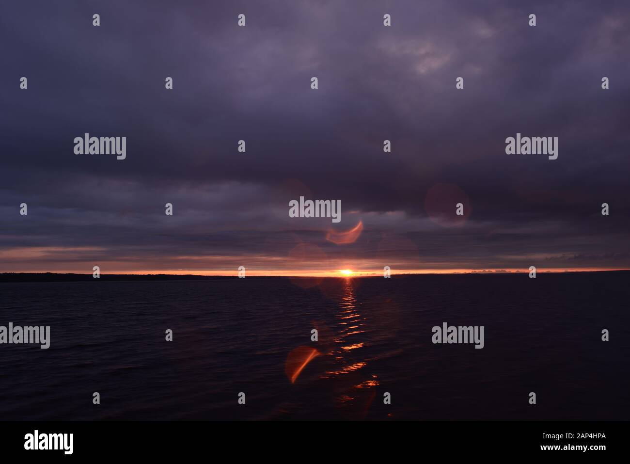 Sonnenstrahl am Himmel am Horizont über der Wasseroberfläche des Sees von unter dunkler Gewitterwolke bei Sonnenuntergang Stockfoto