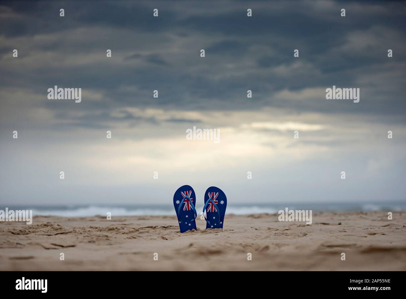 Australische Flaggenthongs, die aufrecht in Sand an einem australischen Strand mit dramatischem Hintergrund von Sturmwolken hängen. Tageskonzept Australien und warten oder beten Stockfoto