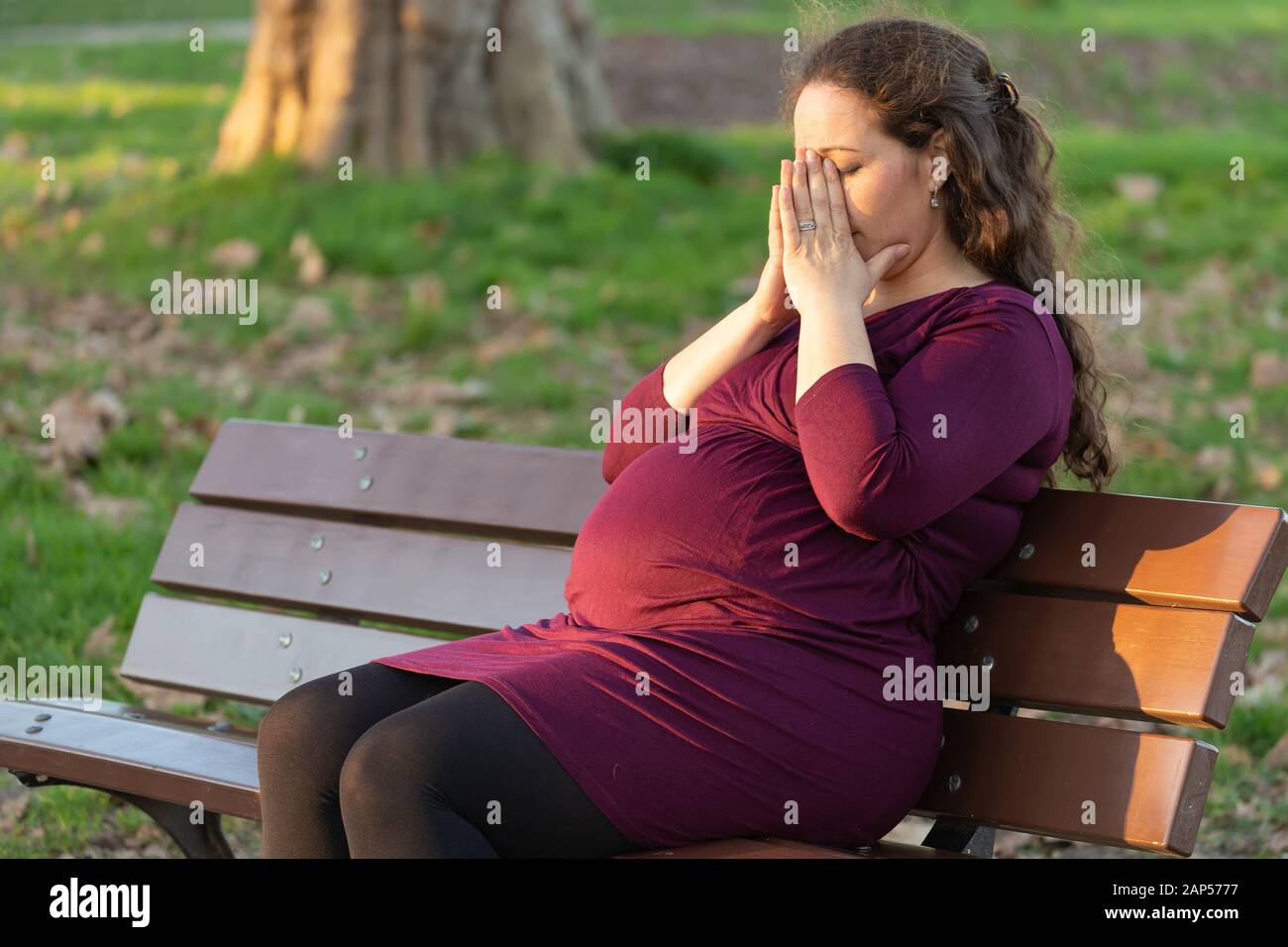 Junge schwangere Frau sich einen Moment Zeit nehmen, zu de-Stress und entspannen Sie sich, wie Sie morgen Krankheit in den warmen Sonnenschein auf einer Parkbank sitzen mit den Händen leidet Cov Stockfoto