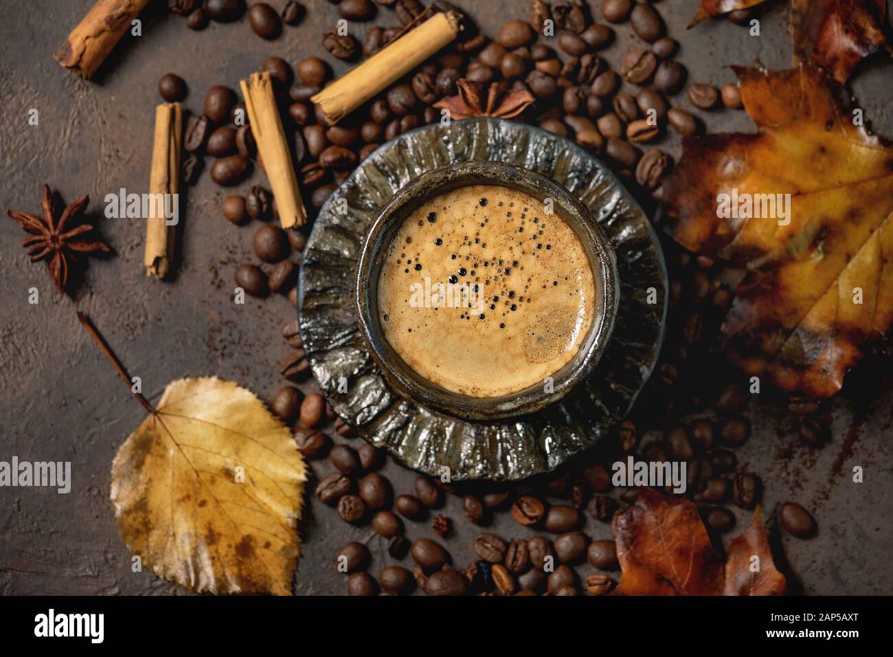Schwarzen Kaffee Espresso mit Schaumstoff in schwarz Keramiktasse mit Untertasse, Herbstblätter, Gewürze und gerösteten Bohnen über braune Textur Hintergrund. Flat l Stockfoto