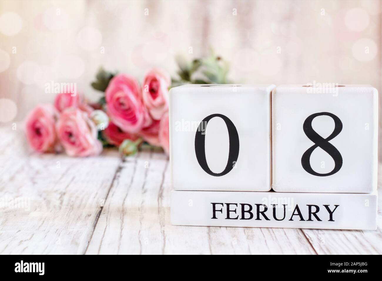 Weiße Holzkalendarkaden mit dem Datum 8. Februar. Selektiver Fokus mit pinkfarbenem Ranunculus im Hintergrund über einem Holztisch. Stockfoto
