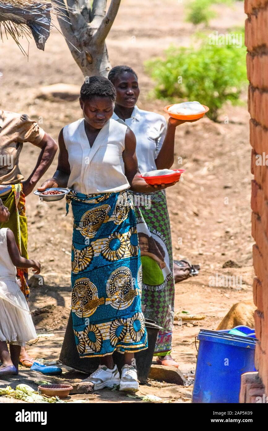 Zwei malawische Frauen tragen Plastikteller und Schalen mit Mais Haferbrei und Bohnen, die ihrer Gemeinschaft serviert werden In einem Dorf in Malaw Stockfoto