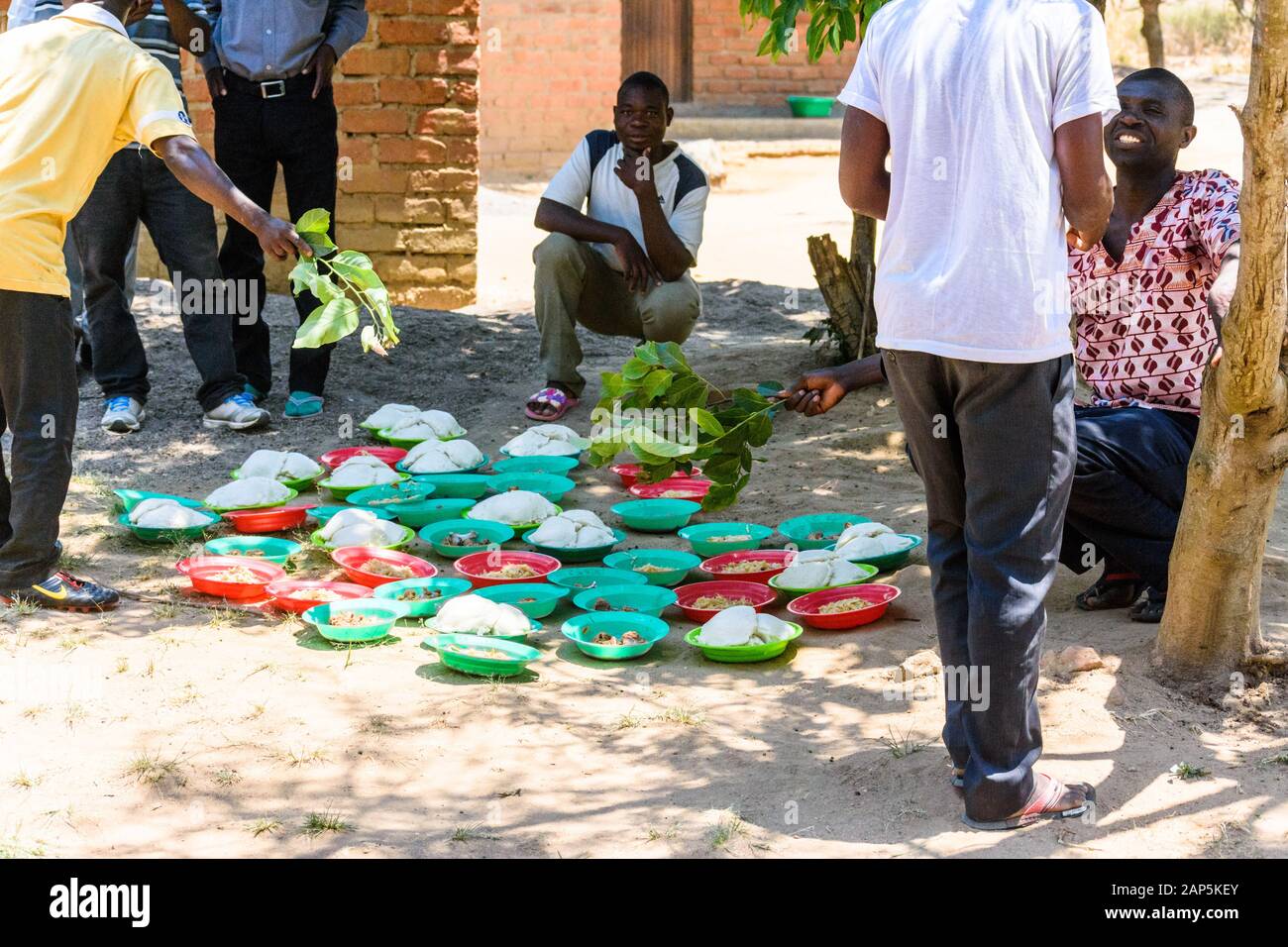 Malawische Männer versammeln sich unter einem Baum, um das Mittagessen von Maisbrei zu essen, der in Plastikschüsseln auf dem Boden aufgestellt ist Stockfoto