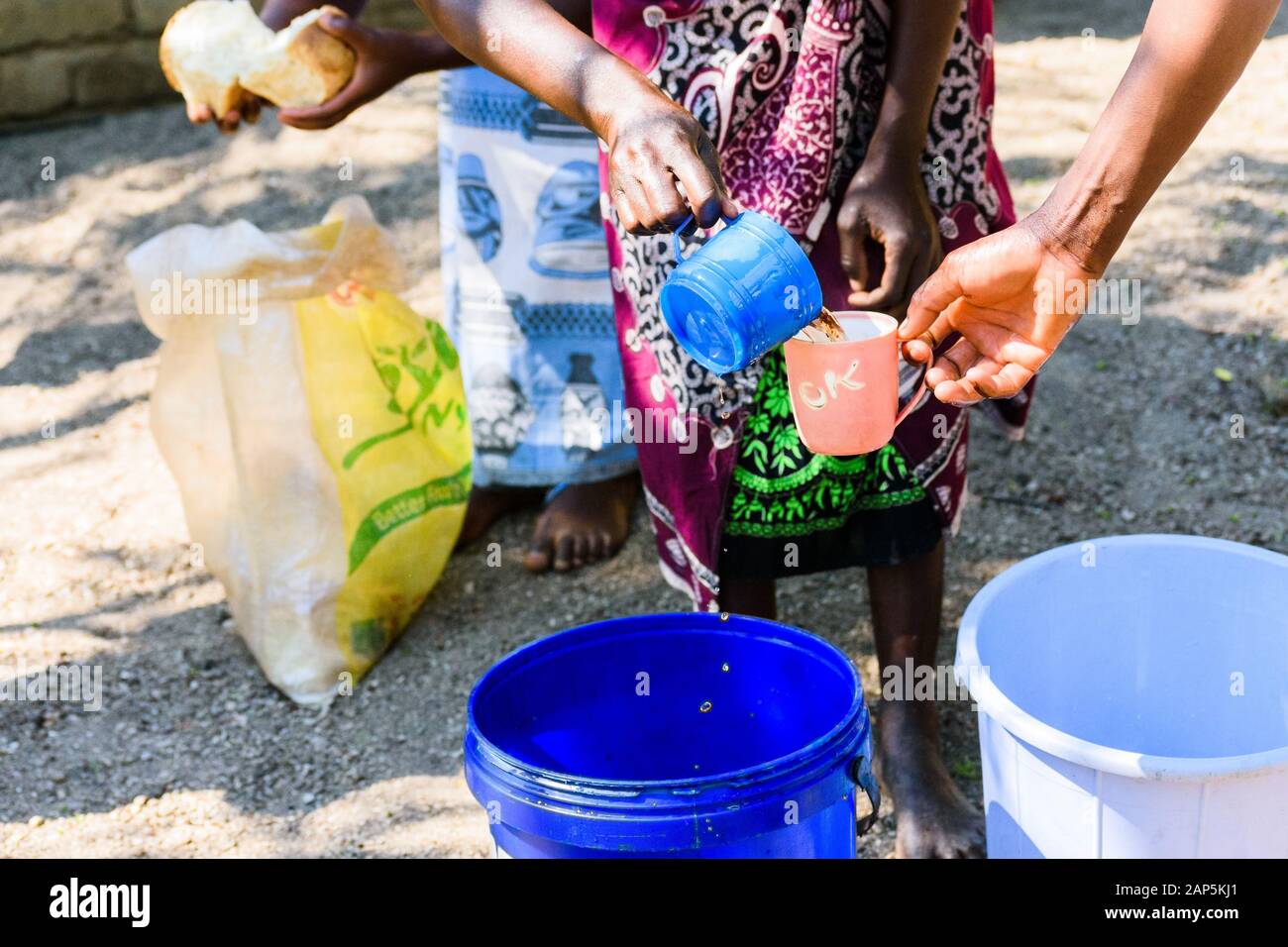Frau gießt Tee in einem großen Plastikeimer gebrüht in Ein Plastikbecher zum Frühstück mit Weißbrot trinken In einem malawischen Dorf Stockfoto