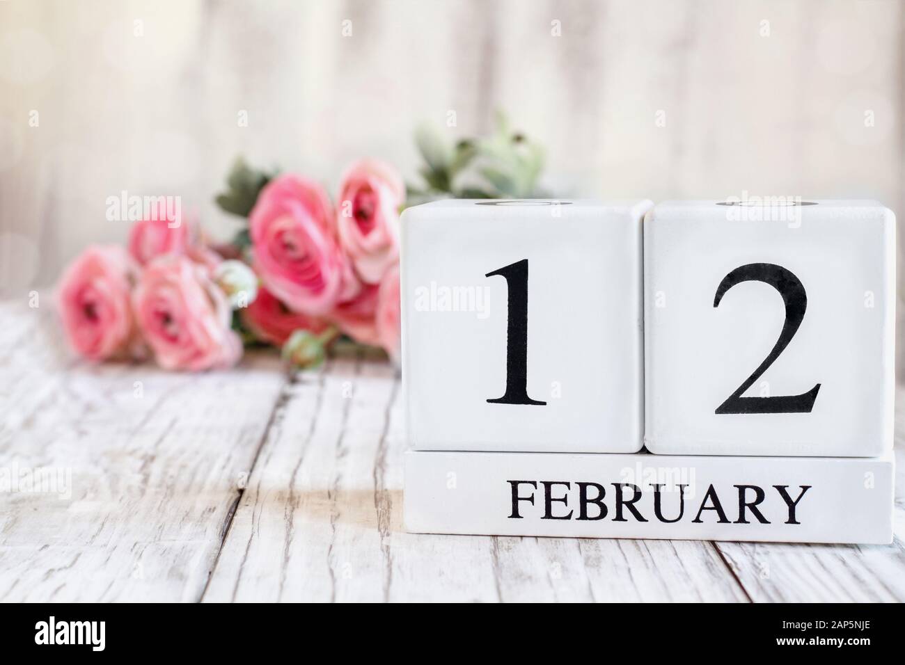 Weiße Holzkalendarkaden mit dem Datum 12. Februar. Selektiver Fokus mit pinkfarbenem Ranunculus im Hintergrund über einem Holztisch. Stockfoto