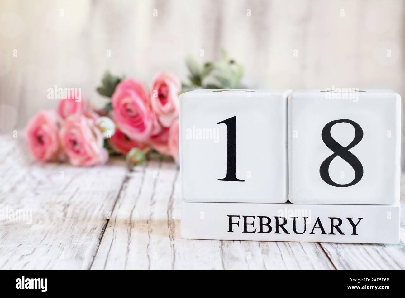 Weiße Holzkalendarkaden mit dem Datum 18. Februar. Selektiver Fokus mit pinkfarbenem Ranunculus im Hintergrund über einem Holztisch. Stockfoto