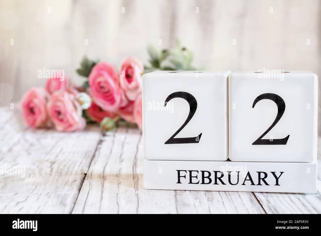 Kalender aus weißem Holz mit dem Datum 22. Februar. Selektiver Fokus mit pinkfarbenem Ranunculus im Hintergrund über einem Holztisch. Stockfoto