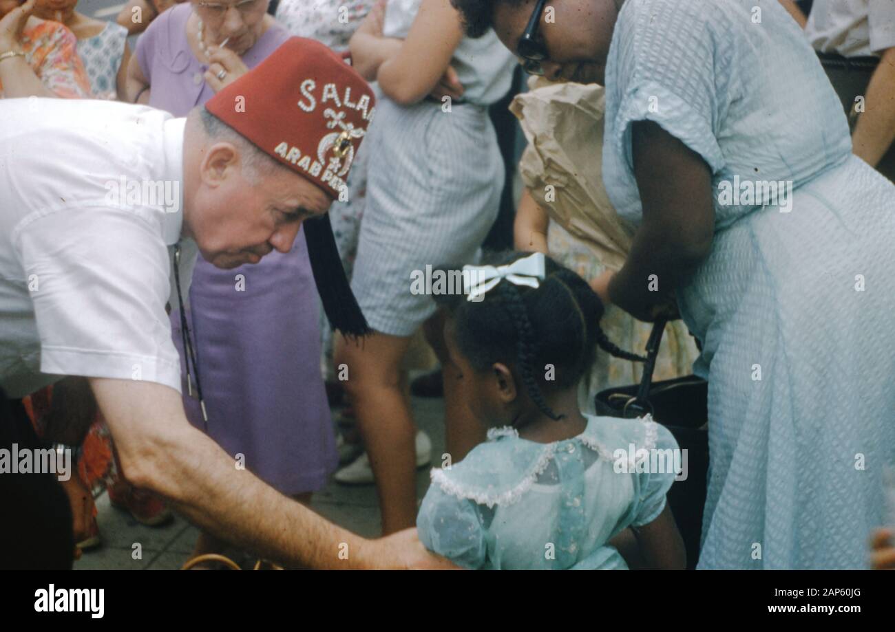 Ein Mann, der einen roten fez-hut mit dem Wort Salam und teilweise sichtbarem Wort Arab trägt, lehnt sich ab, um den Arm eines jungen afroamerikanischen Mädchens in einem blauen Kleid zu halten, das mit einer reifen afroamerikanischen Frau in einer Menschenmenge steht, Vereinigte Staaten, 1965. () Stockfoto