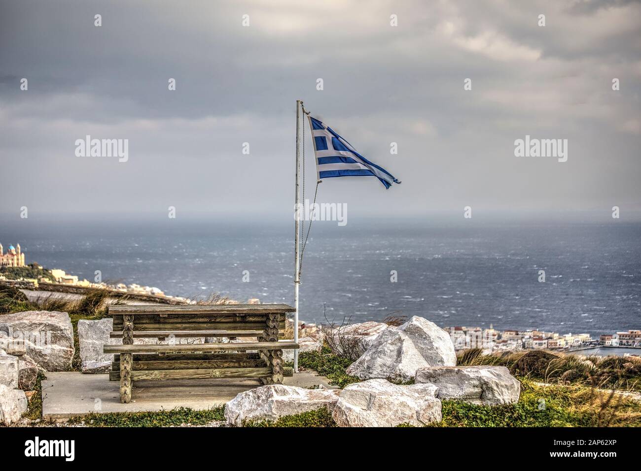 Griechische Flagge flatscht an einem windigen Tag neben dem Picknicktisch aus Holz. Ermoupolis auf der Syros-Insel ganz rechts. Stockfoto