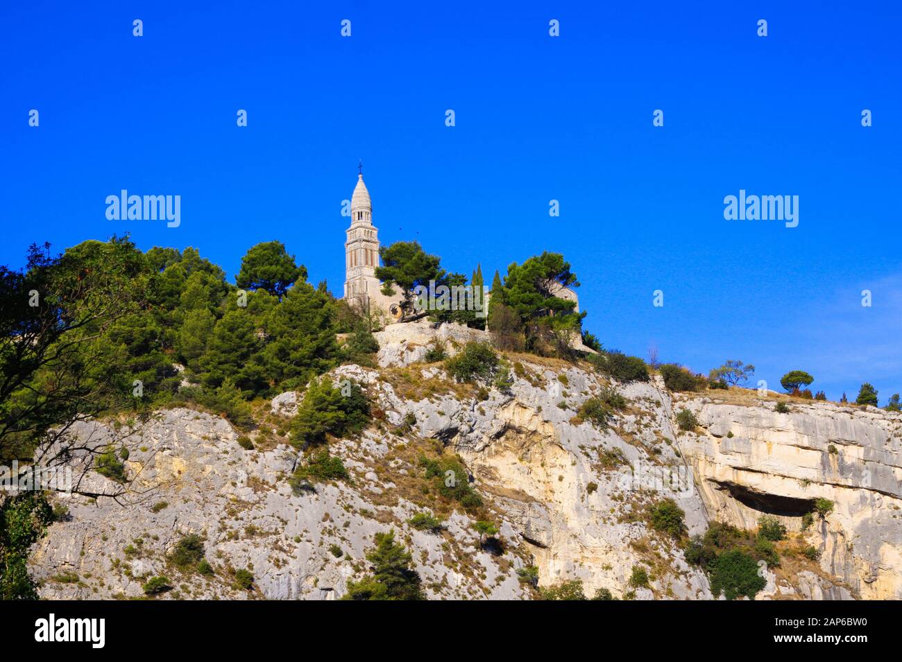 Tiefwinkelansicht auf isolierter römisch-katholischer Kirche auf Berggipfel mit Kiefern- und Zypressbäumen gegen blauen Himmel - Plan d'Orgon, Bouche du Rhône, Nachgewiesen Stockfoto