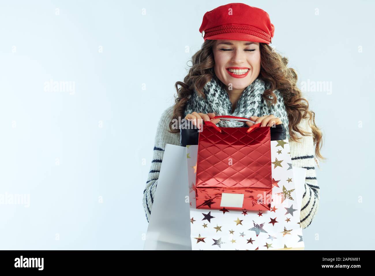 Gerne trendy mittleren Alter Frau mit langen brünetten Haar in Pullover, Schal und Red Hat mit Tüten auf Winter hellblauen Hintergrund isoliert. Stockfoto