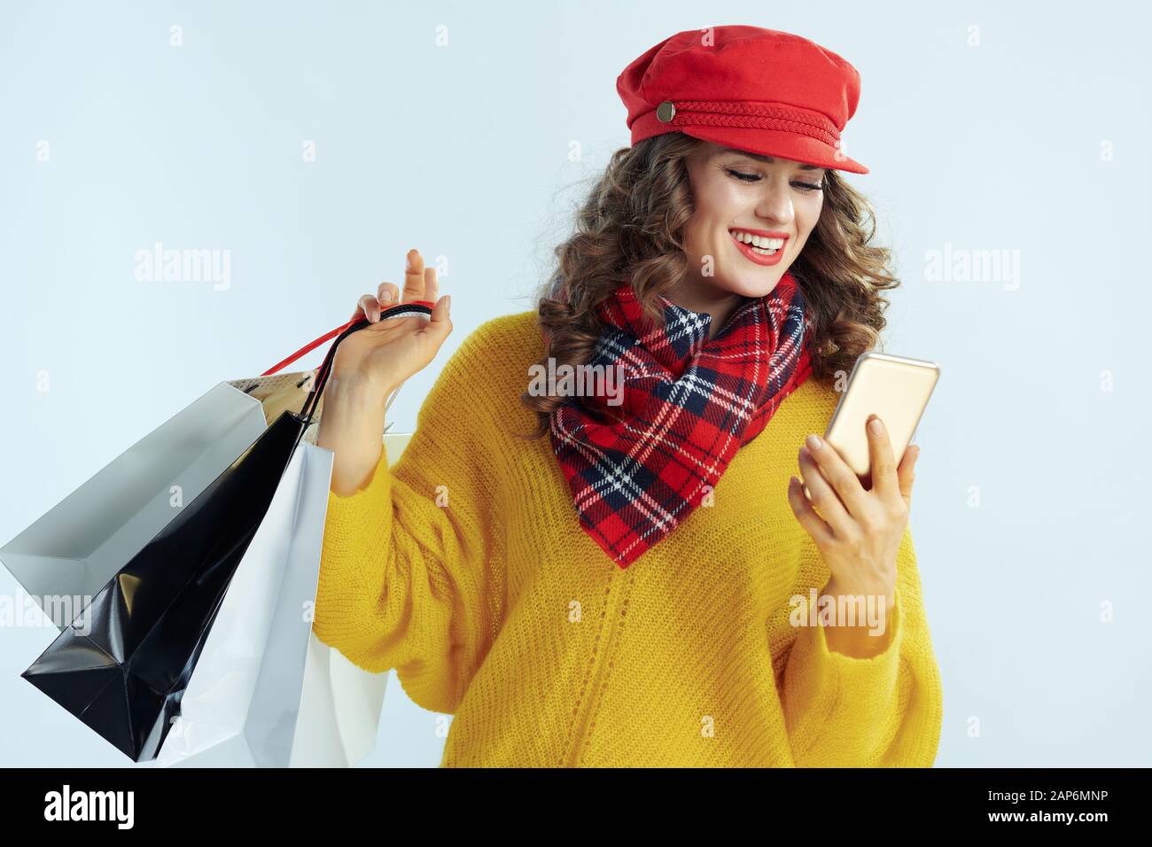 Lächelnd trendy mittleren Alter Frau mit langen brünetten Haar in Pullover, Schal und Red Hat mit Tüten schreiben SMS auf Winter hellblau Bac isoliert Stockfoto
