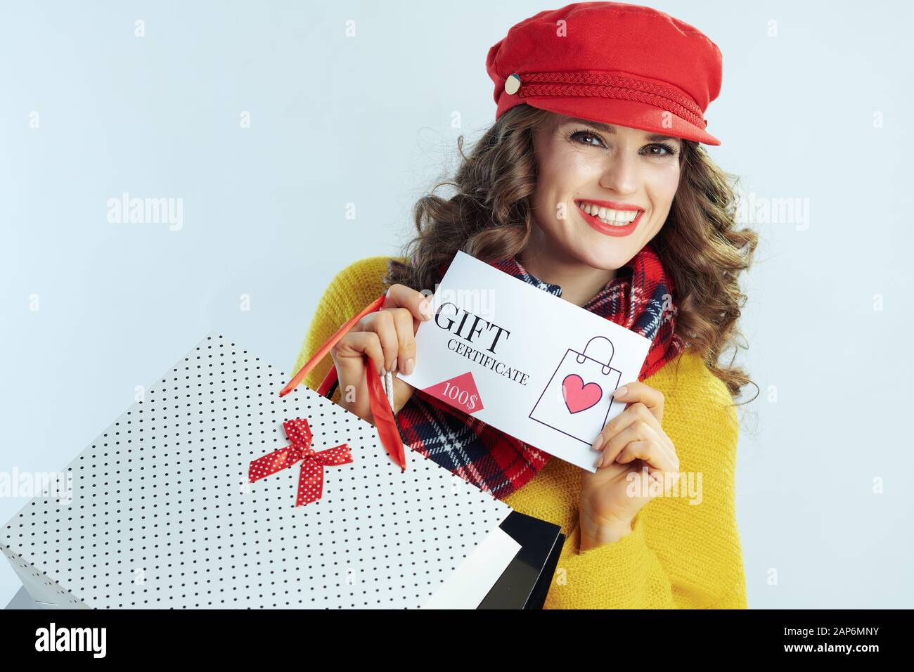 Portrait von Happy elegante 40-jährige Frau mit langen brünetten Haar in Pullover, Schal und Red Hat mit Einkaufstaschen anzeigen Geschenkgutschein gegen Stockfoto