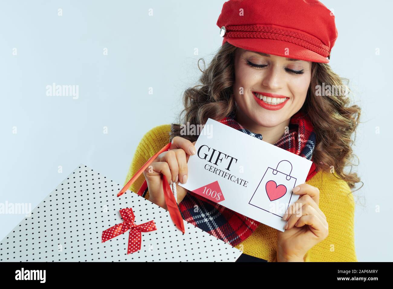 Lächelnd stilvolle Frau mit langen brünetten Haar in Pullover, Schal und Red Hat mit Tüten an Geschenkgutschein gegen Winter Licht, Blau Stockfoto